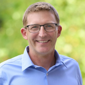 Portrait of an individual smiling, wearing glasses and a blue shirt, set against a blurred green background.