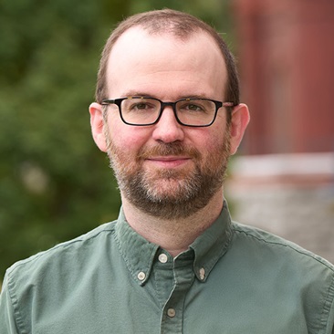 Person with glasses wearing a green shirt, smiling in front of a blurred building background.