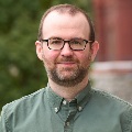 Person with glasses wearing a green shirt, smiling in front of a blurred building background.