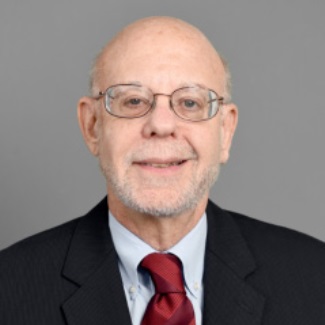 A person with a white beard wearing glasses, a dark suit jacket, white dress shirt, and red tie, photographed against a gray background.
