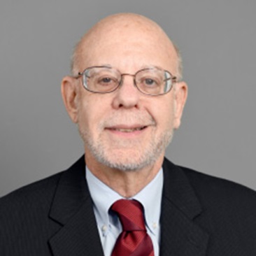 A person with a white beard wearing glasses, a dark suit jacket, white dress shirt, and red tie, photographed against a gray background.