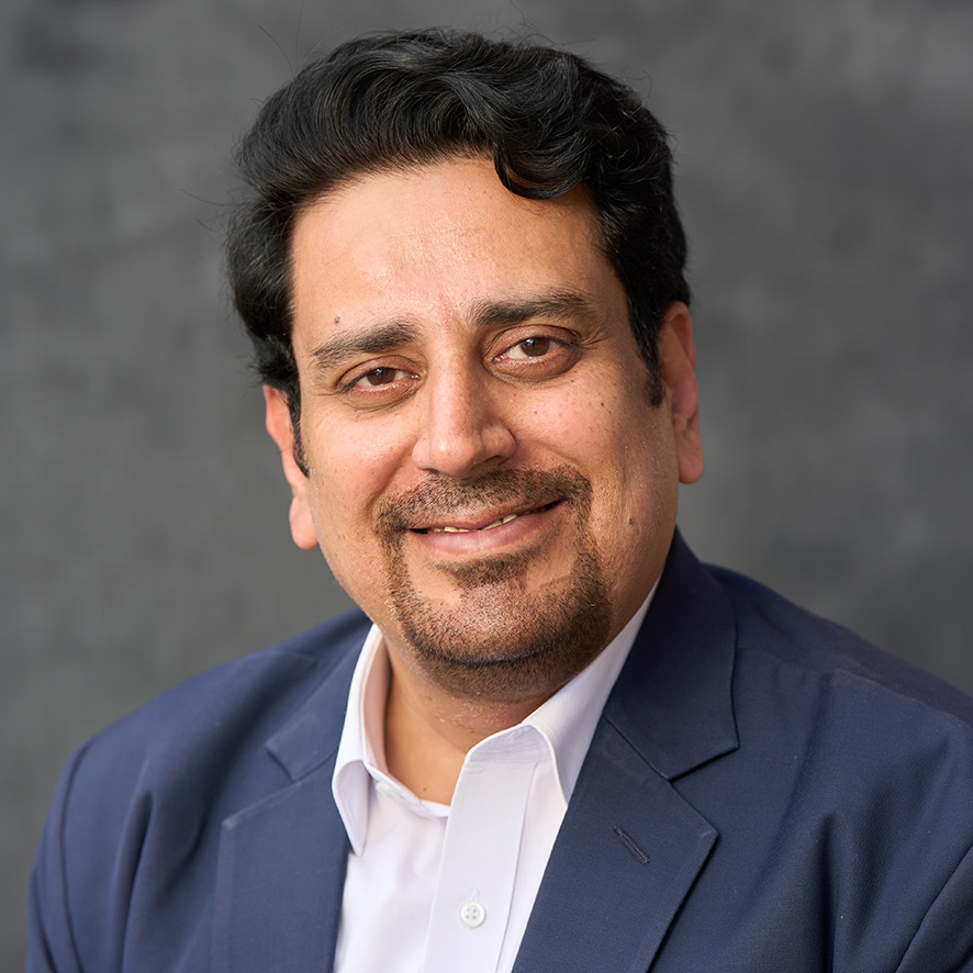 Portrait of a smiling person in a blue suit jacket and light blue shirt, set against a gray background.