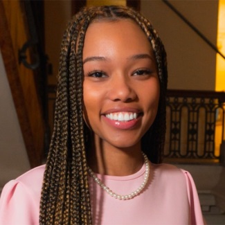Person smiling in a pink outfit and pearl necklace, with long braided hair.