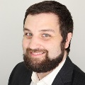 Portrait of a smiling person with a beard, wearing a suit jacket and white shirt.