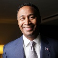 A portrait of a person smiling in a formal blue suit with an American flag lapel pin.