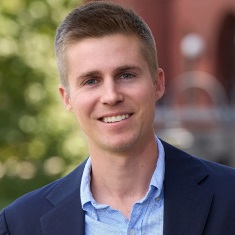 Portrait featuring a smiling individual in a blue suit jacket and patterned shirt, set against a blurred outdoor background.