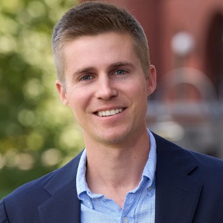 Portrait featuring a smiling individual in a blue suit jacket and patterned shirt, set against a blurred outdoor background.