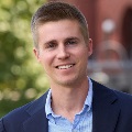 Portrait featuring a smiling individual in a blue suit jacket and patterned shirt, set against a blurred outdoor background.