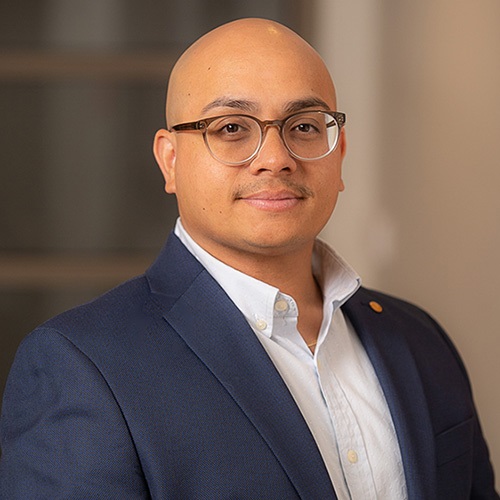 Portrait of a professional in a blue blazer and white shirt, wearing glasses, smiling subtly against a blurred indoor background.