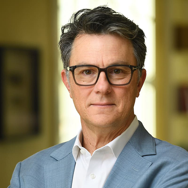 A person with short, dark hair wearing glasses and a white button-down shirt with a light blue blazer.