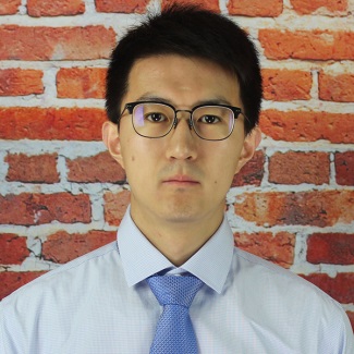 Person with glasses wearing a light blue shirt and blue tie, standing in front of a brick wall.