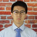 Person with glasses wearing a light blue shirt and blue tie, standing in front of a brick wall.