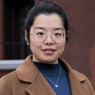 Smiling person with glasses, camel coat, and blue sweater in front of a brick building.