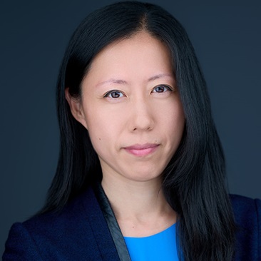 A person with long straight black hair, wearing a bright blue top and navy blazer, against a dark gray background.