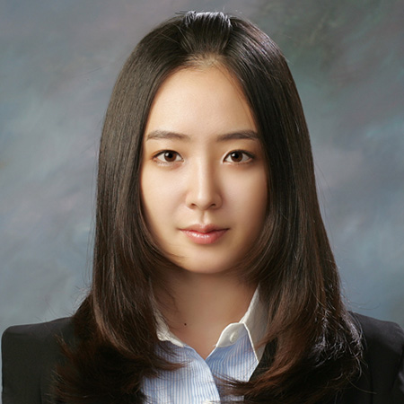 A person wearing a black blazer over a light blue collared shirt, set against a muted gray-blue background.