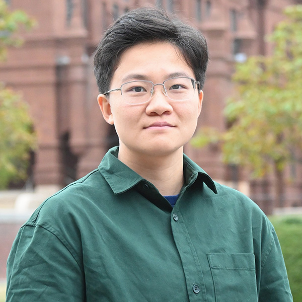 A person in a green shirt and glasses, standing outdoors in front of a brick building.