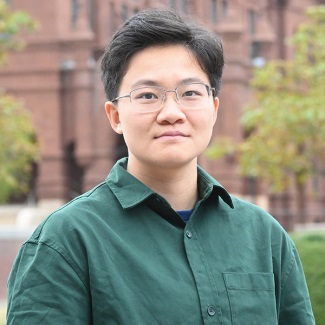 A person in a green shirt and glasses, standing outdoors in front of a brick building.