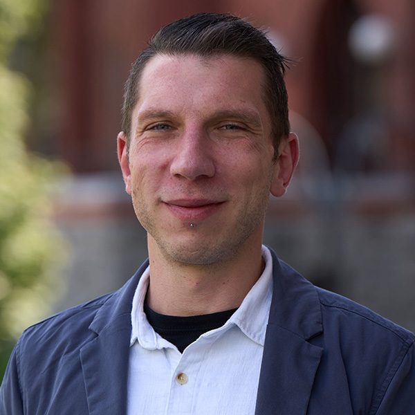 Portrait of a smiling person with short hair, wearing a black jacket over a white collared shirt, standing in front of a brick building.