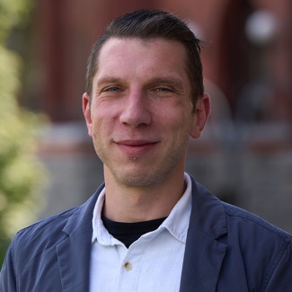 Portrait of a smiling person with short hair, wearing a black jacket over a white collared shirt, standing in front of a brick building.