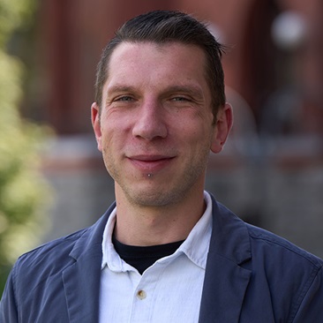Portrait of a smiling person with short hair, wearing a black jacket over a white collared shirt, standing in front of a brick building.