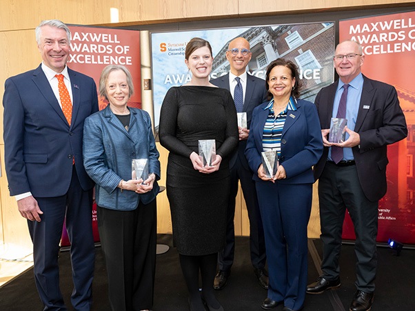A group of six individuals is standing indoors, each holding a small award. Behind them, banners read "Maxwell Awards of Excellence" and display the Syracuse University logo. They are dressed in formal attire and are smiling at the camera.