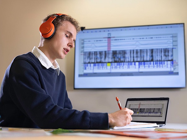 A person wearing orange headphones is sitting at a desk, writing on paper. In front of them is a laptop showing a waveform or audio editing software. A larger screen in the background displays a similar interface.