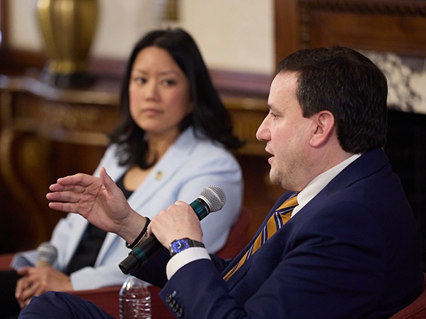 Two individuals are speaking on a panel. One person is gesturing while holding a microphone. They're seated in a room with ornate furniture and a decorative lamp.