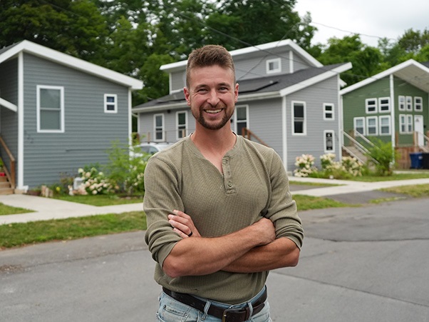 A person stands with arms crossed on a street lined with tiny homes.
