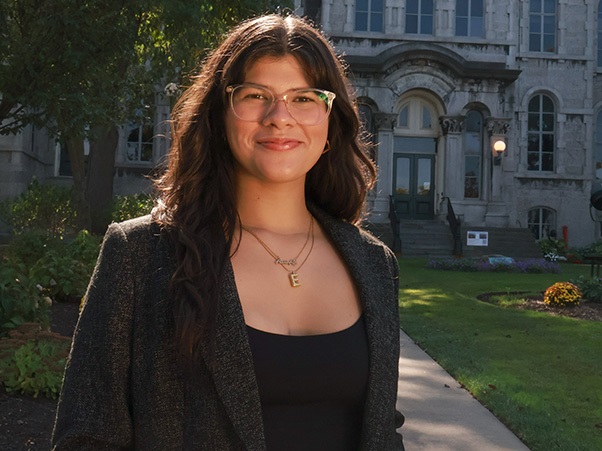 A person with long dark wavy hair and glasses, wearing a black top and gray tweed blazer with a gold necklace, smiling while standing outdoors in front of a stone campus building with a green lawn.