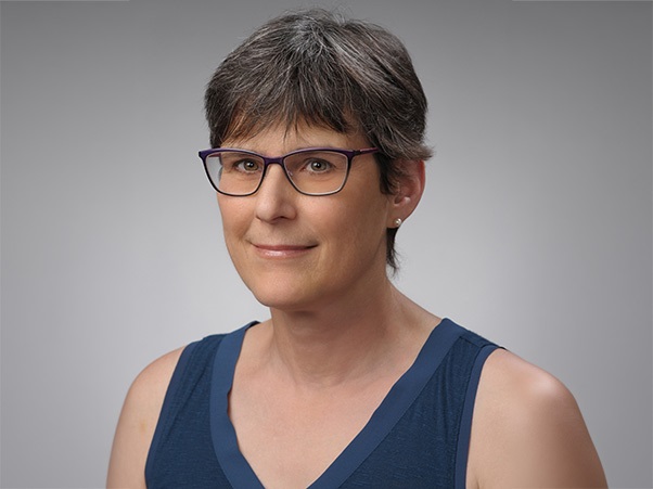 A person with short salt-and-pepper hair and purple-framed glasses, wearing a sleeveless navy blue v-neck top, against a gray studio background.