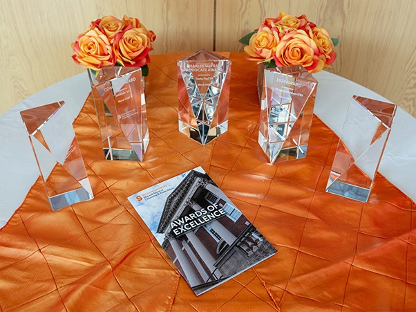 A table with an orange cloth holds five crystal awards and two bouquets of orange roses. In the center is a pamphlet titled "Awards of Excellence."