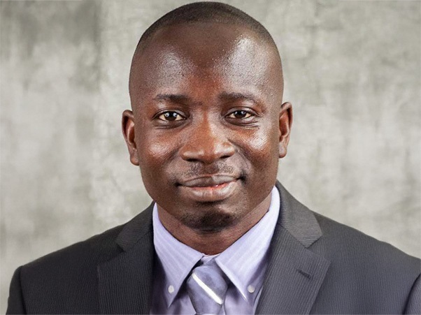 A person with a subtle smile, wearing a dark suit and tie against a textured gray background.
