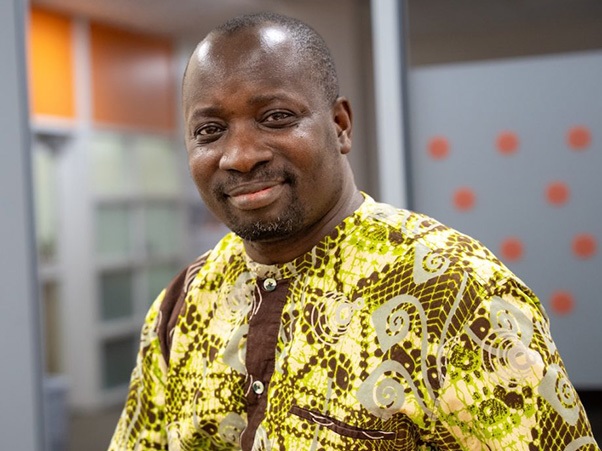 A person smiling, wearing a patterned yellow and brown traditional shirt.