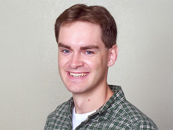 Smiling person with short brown hair and a green plaid collar in front of a neutral backdrop.
