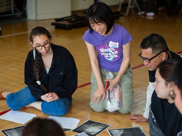 A group of people sitting on the floor in a classroom, engaged in discussing black-and-white photographs spread out on the ground. One person is holding a set of papers and smiling. The environment appears to be informal and collaborative.