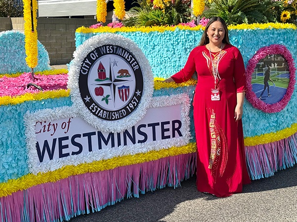 A person stands next to a colorful float decorated with flowers and a sign that reads "City of Westminster, Established 1957." The float features the city's emblem and the person is wearing a red outfit with a name badge.