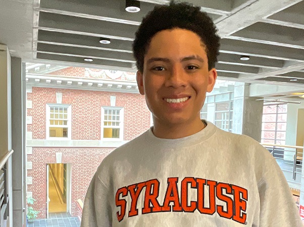 A person smiles at the camera wearing a gray Syracuse University sweatshirt. A brick building is visible through large windows in the background.