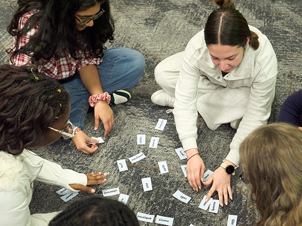 A group of people sitting on a carpeted floor, engaged in a word card activity. The cards are scattered in front of them, and each person is reaching for different cards.