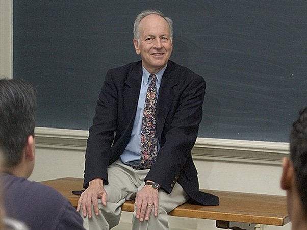 Generate by AI Assist  A person sits on a desk in front of a chalkboard, smiling and facing an audience. The individual is wearing a dark blazer and patterned tie.