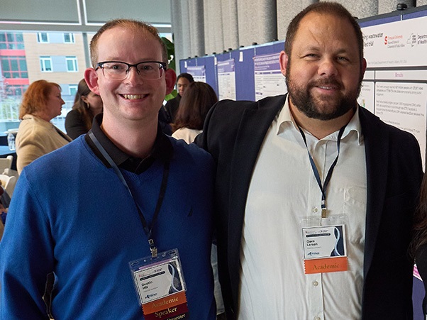 Two people stand together and smile for a photo at an academic conference, with research poster displays and attendees visible in the background. Both wear conference lanyards; one wears a blue sweater and glasses, the other a dark blazer.