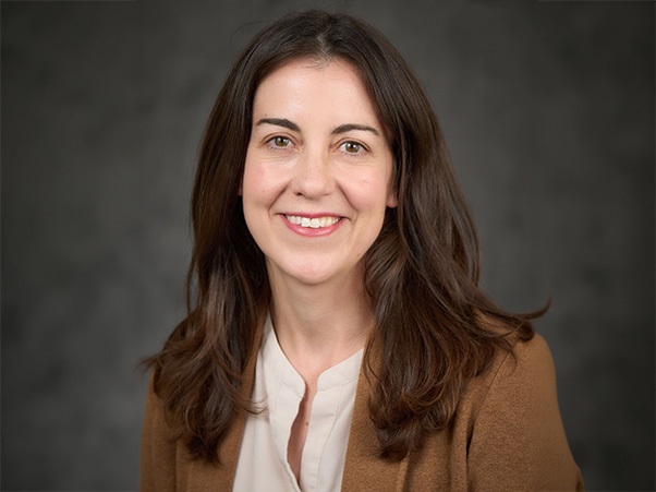 A person smiling, long brown hair, wearing a cream top and brown cardigan, gray backdrop.
