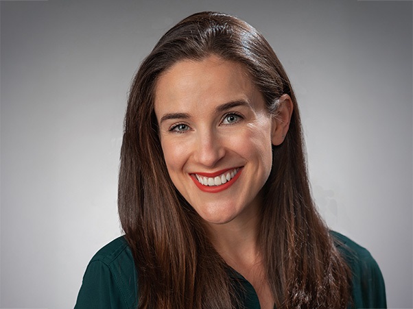 A person smiling, with long brown hair, wearing a teal top, against a gray background.