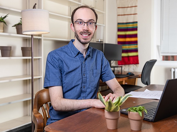 A person sits at a wooden desk with a laptop and two small potted plants, smiling at the camera. They are wearing glasses and a blue short-sleeve button-down shirt. Bookshelves, a floor lamp, and a colorful woven wall hanging are visible.
