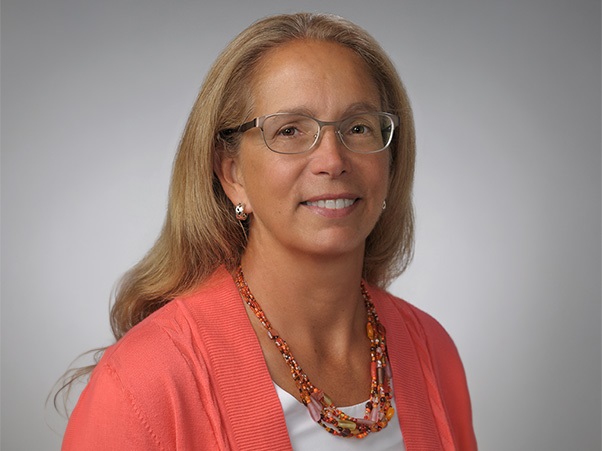 A person with shoulder-length blonde hair wearing glasses, a coral cardigan, and a beaded necklace in orange and amber tones, against a light gray background.