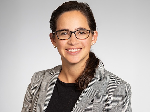 A person with dark hair pulled back, wearing tortoiseshell glasses, pearl stud earrings, and a plaid blazer, smiling at the camera against a light gray background.