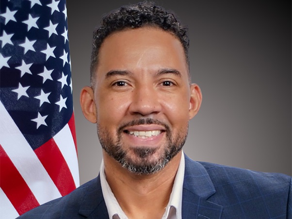 A person smiling wearing a dark suit. There is an American flag in the background.