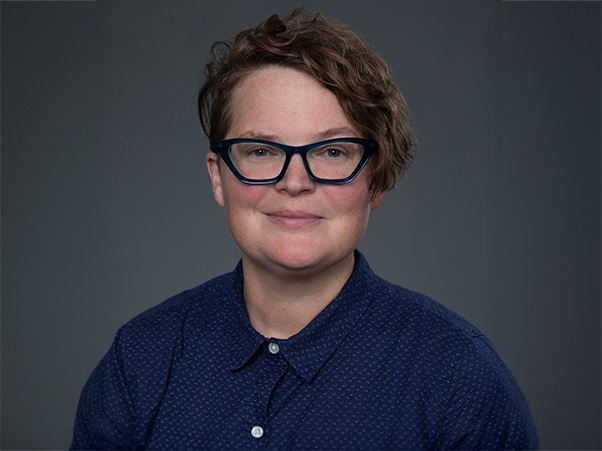 A person with short brown hair wearing dark glasses and a dark button down shirt.