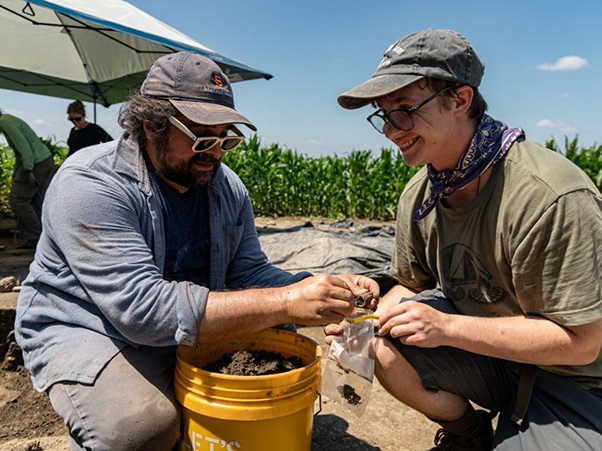 Two people crouch over a bucket, examining soil samples at an outdoor excavation site.