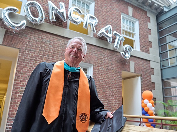 A person in graduation attire stands outside a building with the word "CONGRATS" spelled in balloon letters above. The person holds a cap and smiles at the camera. Orange and blue balloons are visible in the background.