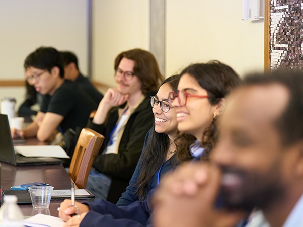 A group of people sitting at a conference table, engaged in discussion. Some are using laptops and taking notes. They appear attentive and focused.